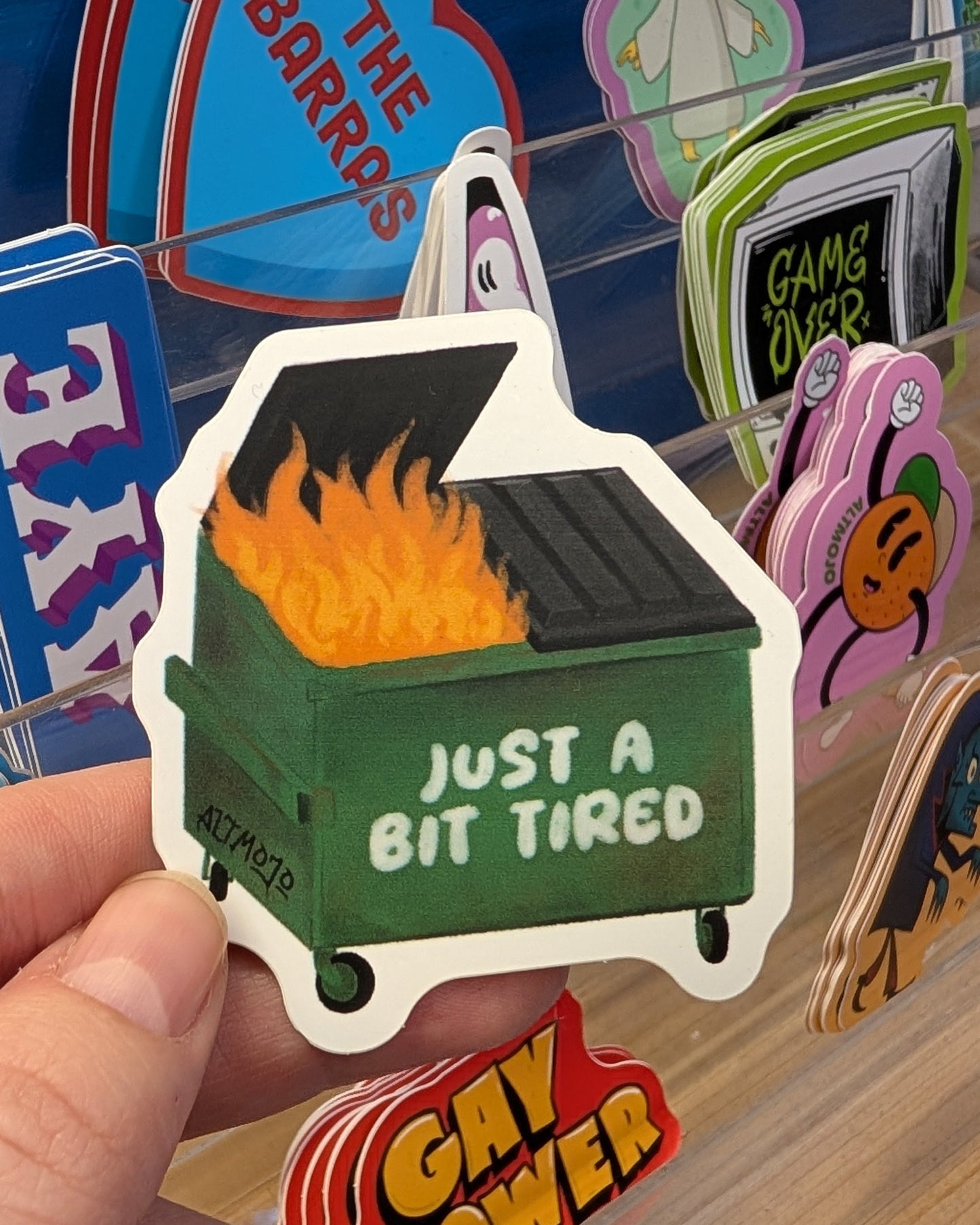 Hand holding a sticker of a green dumpster with flames and 'Just a Bit Tired' text, with a colorful background of other stickers.