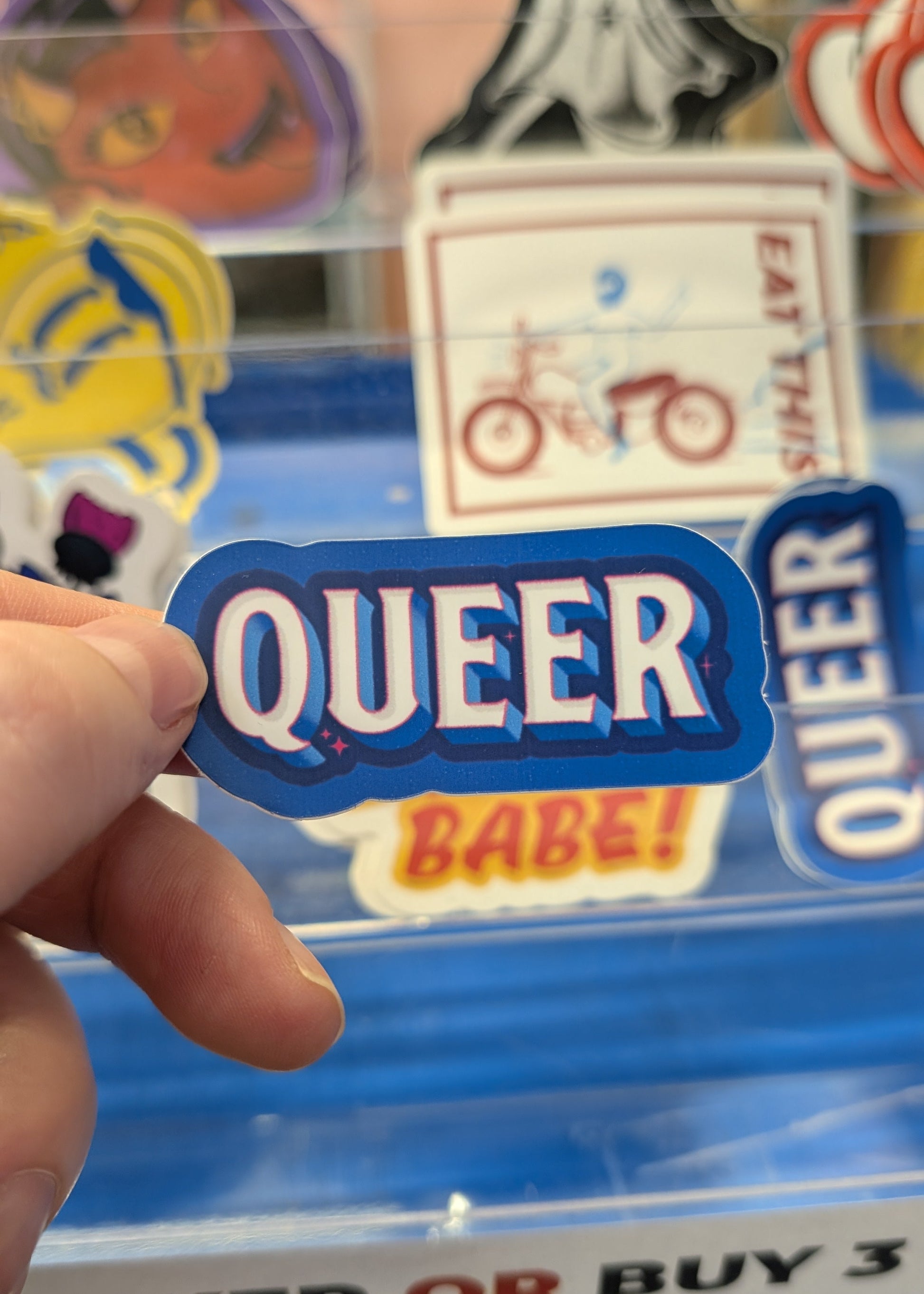 Hand holding a 'QUEER' sticker in front of a display with other stickers.