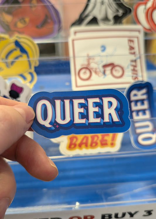 Hand holding a 'QUEER' sticker in front of a display with other stickers.