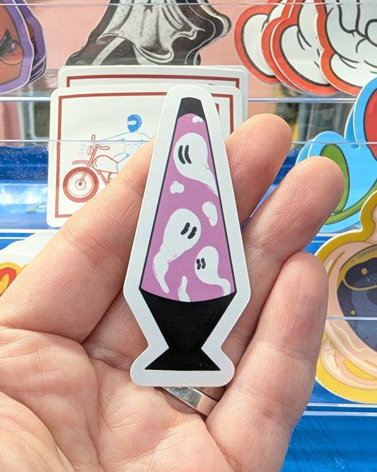 Hand holding a sticker of a pink lava lamp with ghost design against a colorful background